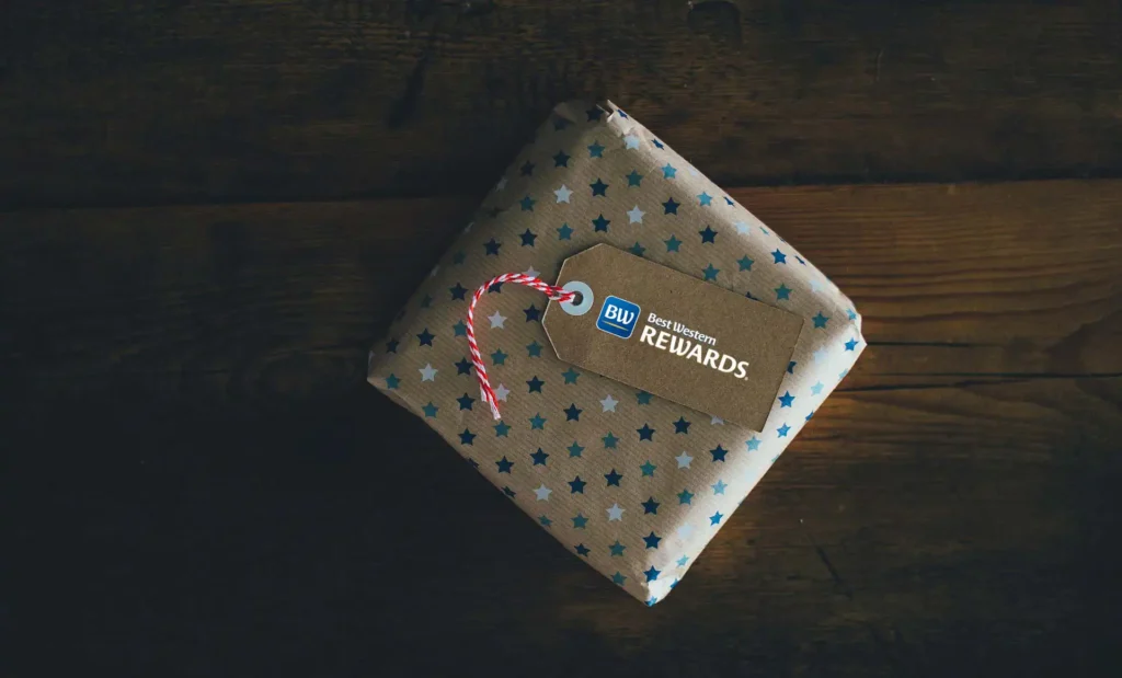 In the image a gift package is placed on a wooden table. The package is brown with blue and light blue stars. On top is a brown tag with the Best Western Rewards® logo