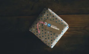 In the image a gift package is placed on a wooden table. The package is brown with blue and light blue stars. On top is a brown tag with the Best Western Rewards® logo