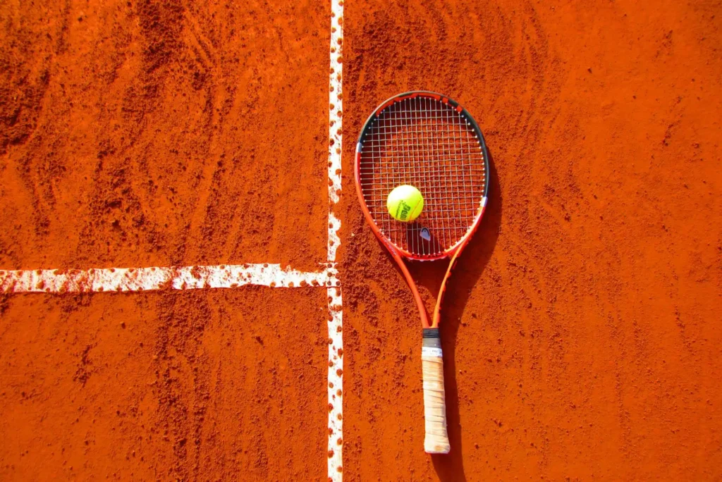 Nell'immagine una racchetta da tennis posata su un campo. Sopra la racchetta è posata una pallina da tennis.